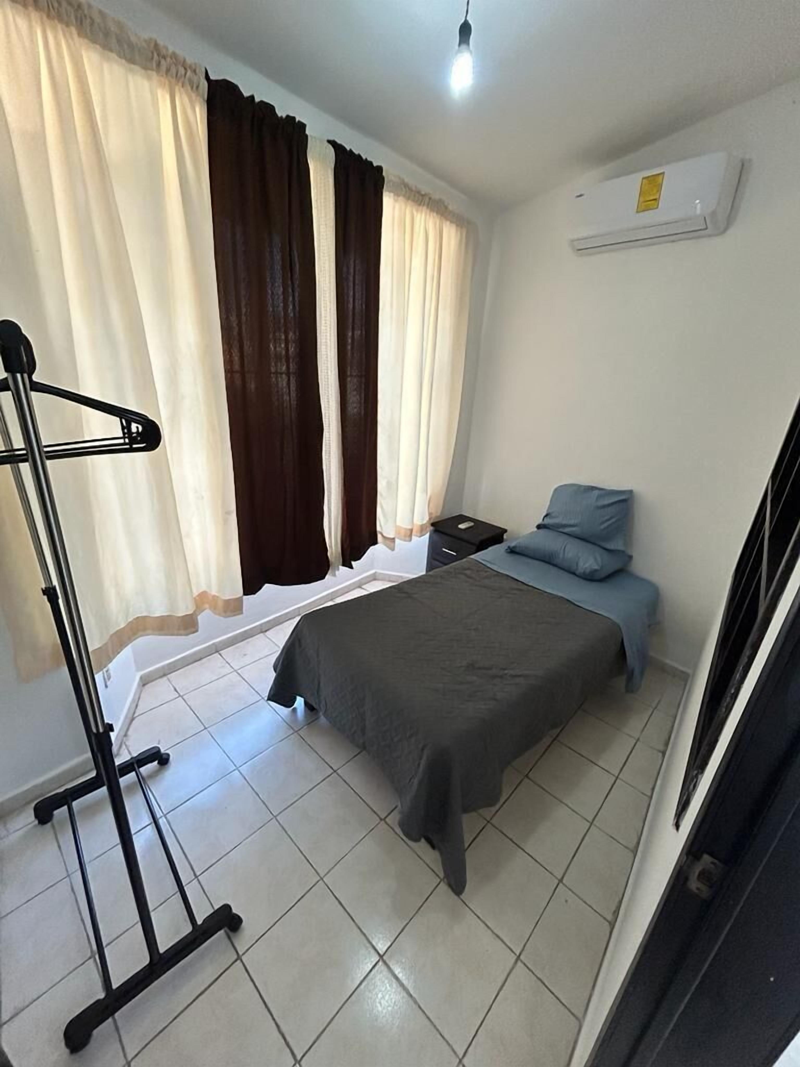 Un dormitorio con cama, mesita de noche, escritorio, silla, lámpara colgante y ventana con cortinas.