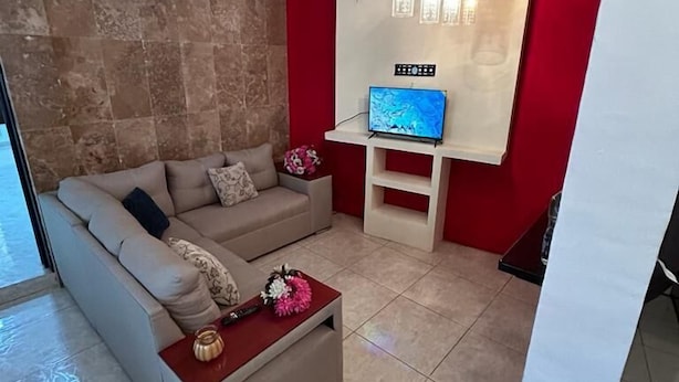Uma sala de estar moderna com um sofá bege, uma mesa de centro com flores, uma TV de tela plana fixada em uma parede vermelha e um lustre.