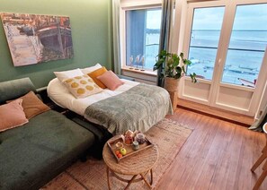 Ein Schlafzimmer mit einem Bett, zwei Sofas, einem kleinen Tisch mit einer Auflage und Gegenständen, einem großen Fenster mit Meerblick und einem Gemälde an der Wand.