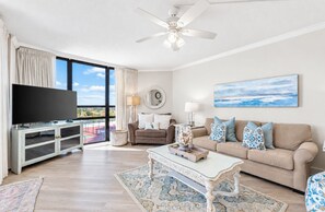 A spacious living room with a flat-screen TV, a ceiling fan, a large window with curtains, a beige sofa with patterned cushions, and a decorative table. - Gulf views+ Private beach! Restaurant on site! (Santa Rosa Beach)