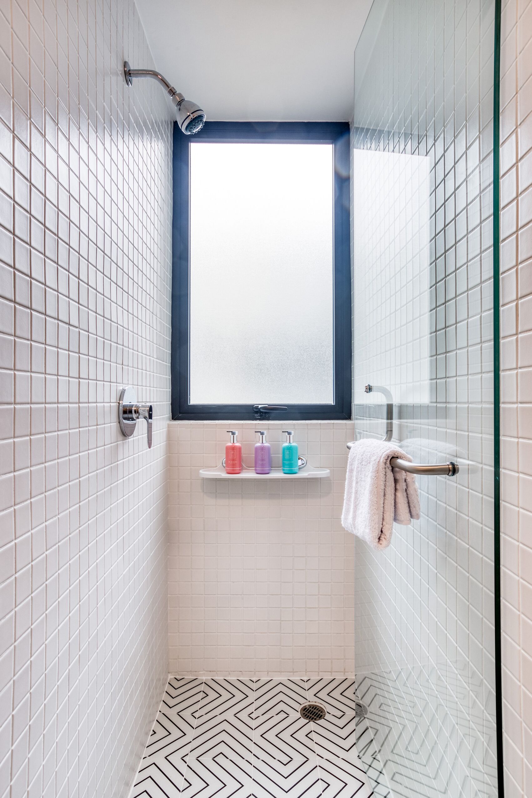 A small, modern bathroom with white tiled walls, a shower with a handheld showerhead, a window, and a shelf with toiletries.