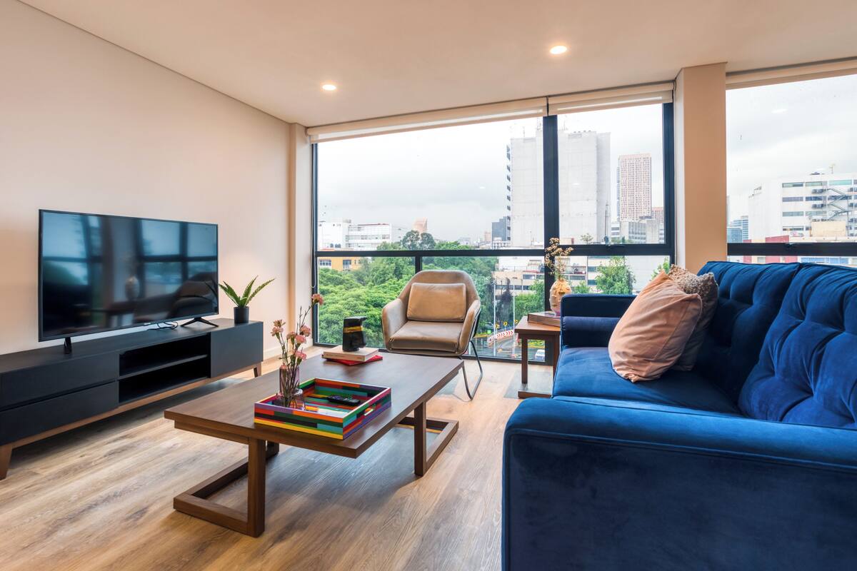 Un salon moderne doté d’un grand téléviseur, d’un canapé bleu, d’une table basse et offrant une vue sur la ville grâce à de grandes fenêtres.