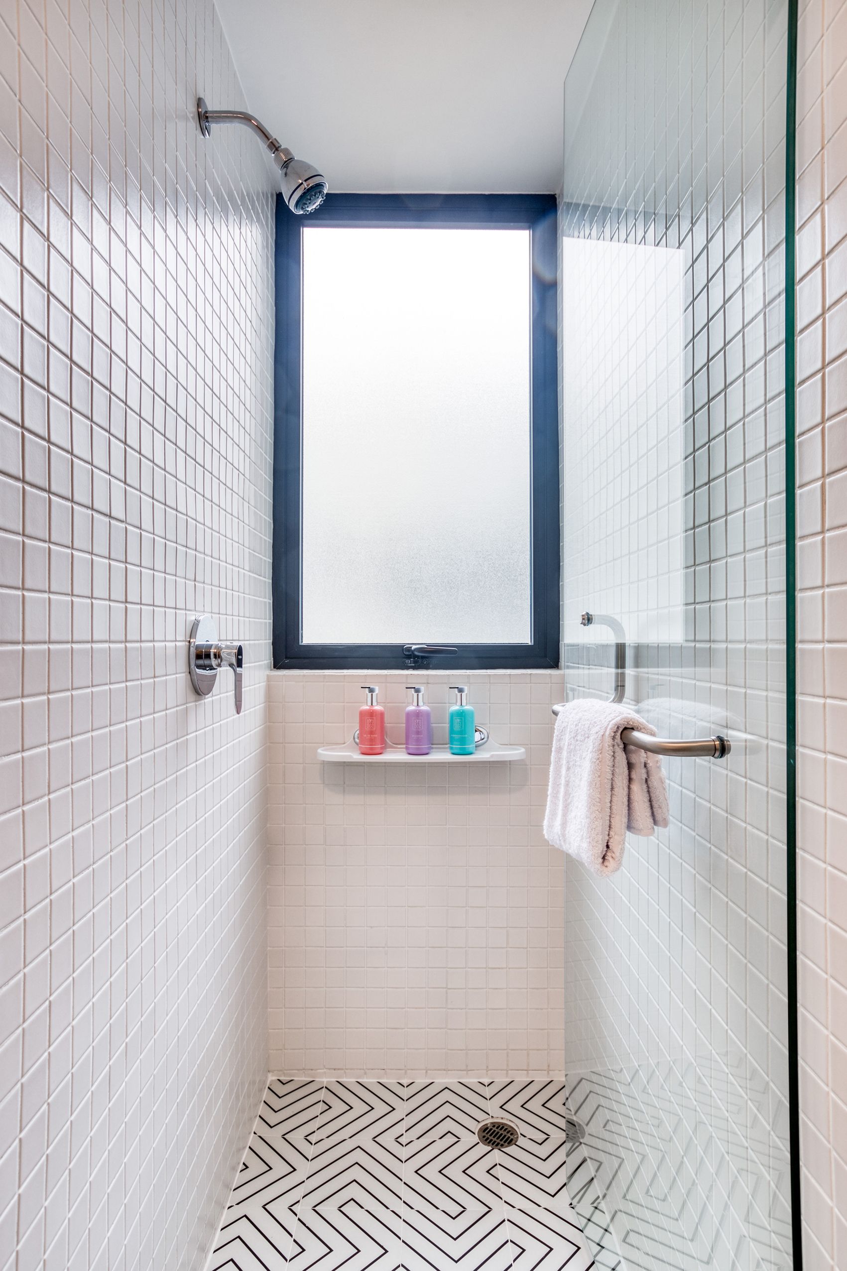 Une petite salle de bain moderne avec des murs recouverts de carreaux blancs, une douche avec un pommeau de douche amovible, une fenêtre et une étagère contenant des articles de toilette.