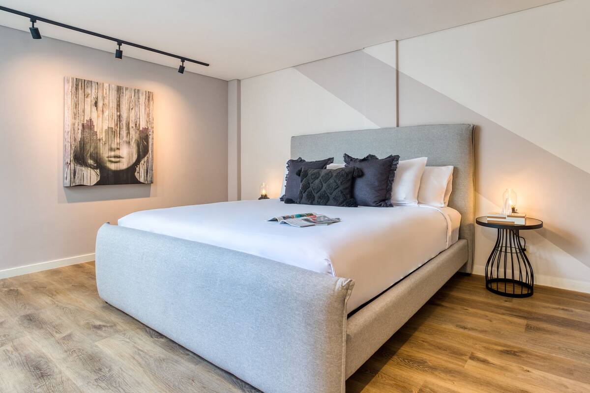 Une chambre à coucher moderne avec un grand lit, une table de chevet avec une lampe et une œuvre d’art représentant un paysage urbain accrochée au mur.