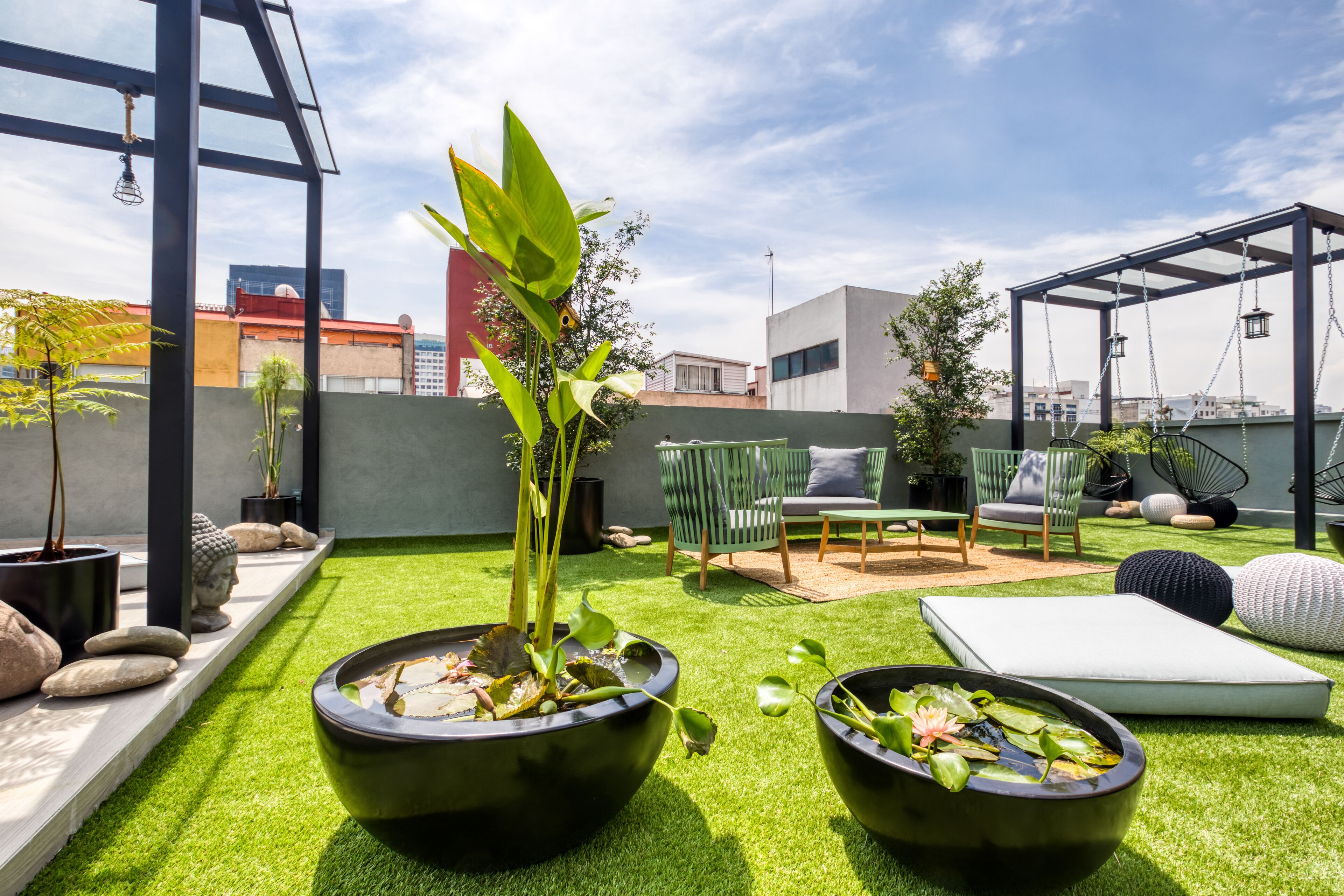 A rooftop garden with artificial grass, two large black planters with water lilies, a green chair, a small table, and a pergola with hanging lights.