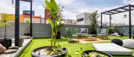 A rooftop garden with artificial grass, two large black planters with water lilies, a green chair, a small table, and a pergola with hanging lights.