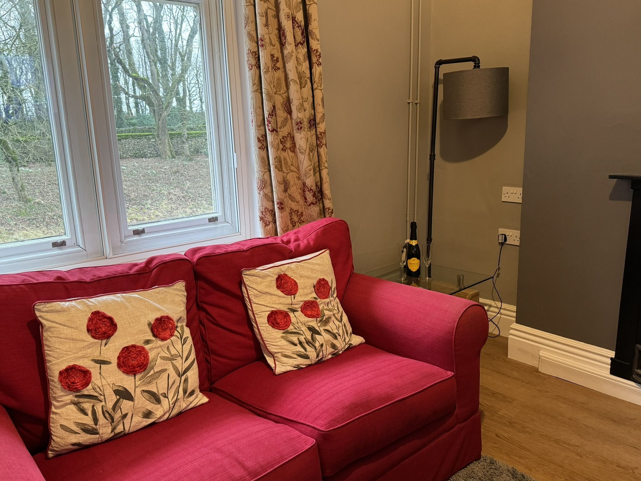 A red sofa with floral cushions, a floor lamp, and a window with curtains.