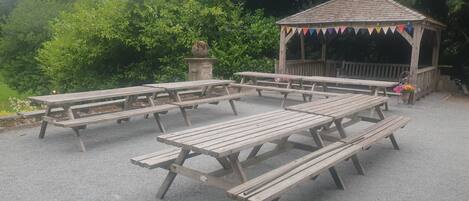 A gazebo with picnic tables arranged around it, surrounded by greenery.