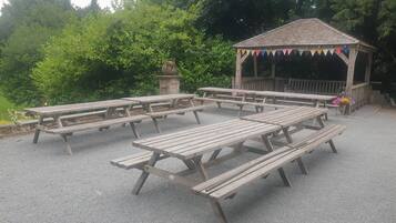 A gazebo with picnic tables arranged around it, surrounded by greenery.