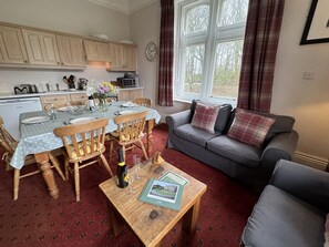 A cozy living room with a grey sofa, wooden dining table set for two, and a red patterned carpet.