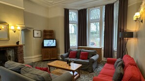 A living room with a fireplace, a television, and a large window with curtains. - Cosy self catering apartment in large Victorian Residence (Buxton)