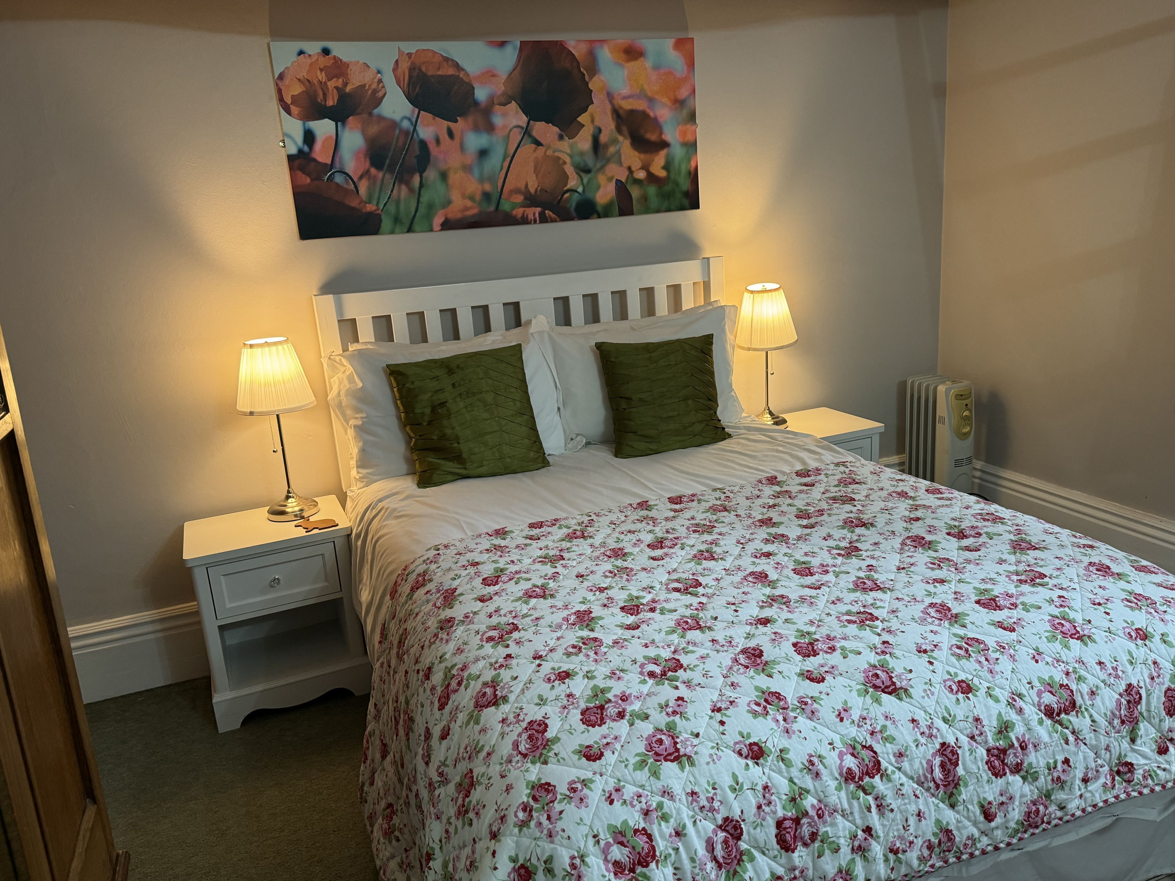 Ein ordentlich bezogenes Bett mit floralem Bettzeug, zwei Nachttischlampen und ein Blumenbild an der Wand.