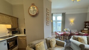 Un salon confortable avec deux canapés, un coin repas avec une table dressée pour quatre personnes, une cuisine équipée d’un four à micro-ondes et d’un four traditionnel, et une grande horloge murale.