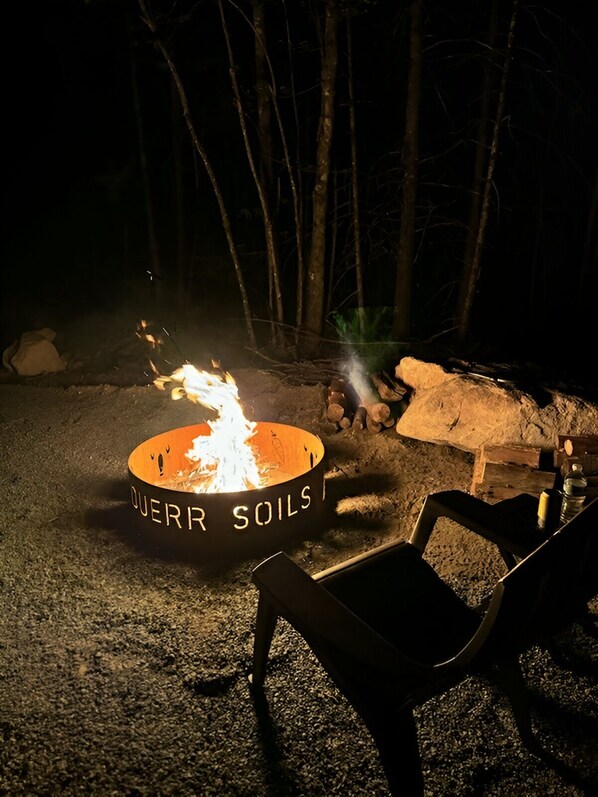 A campfire with a metal bowl labeled "QUERR SOILS" surrounded by rocks and logs.