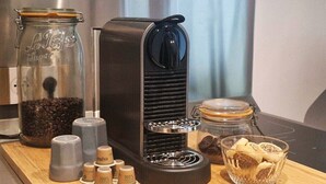 A coffee maker, a glass jar with coffee beans, and a glass container with coffee pods on a wooden surface.