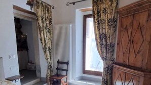 A bedroom with a large bed, wooden headboard, a wooden wardrobe, and a chair. There is a window with curtains and a hanging light fixture.