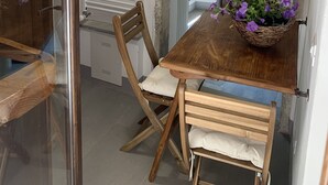 A dining area with a wooden table, chairs, a basket of flowers, and a shelf with kitchen items.