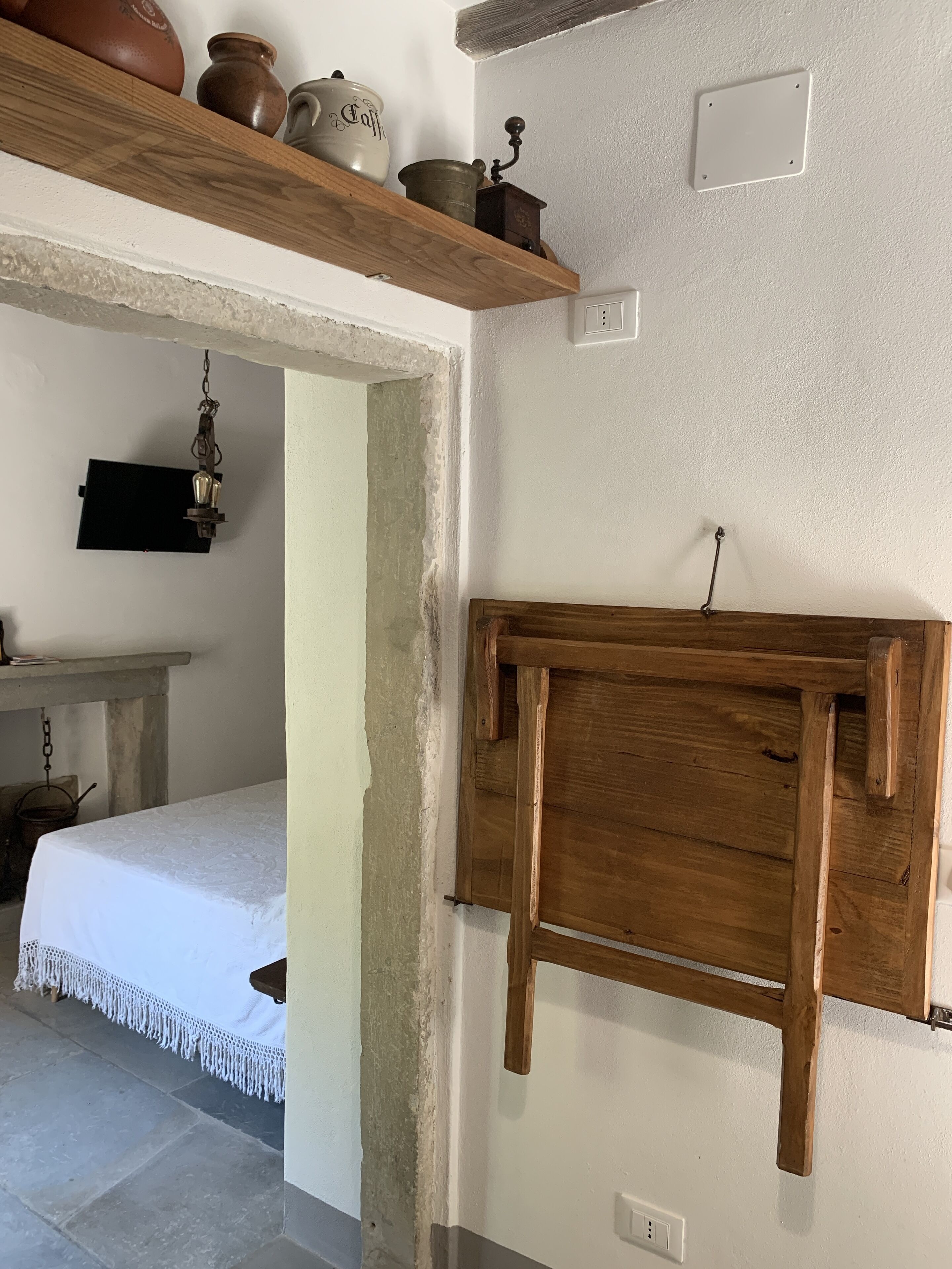Een eenvoudige kamer met een houten bed, een klein tafeltje en een plank met keramiek.