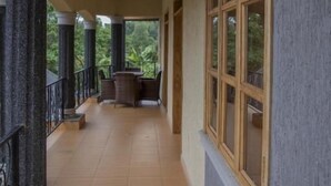 A covered corridor with wooden beams and columns, a seating area with chairs, and large windows offering a view of greenery. - Mt. Rwenzori Golf Resort & Spa (Fort Portal)