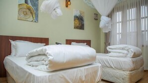 A hotel room with two beds, a wooden headboard, a large window with sheer curtains, and wall art. - Mt. Rwenzori Golf Resort & Spa (Fort Portal)