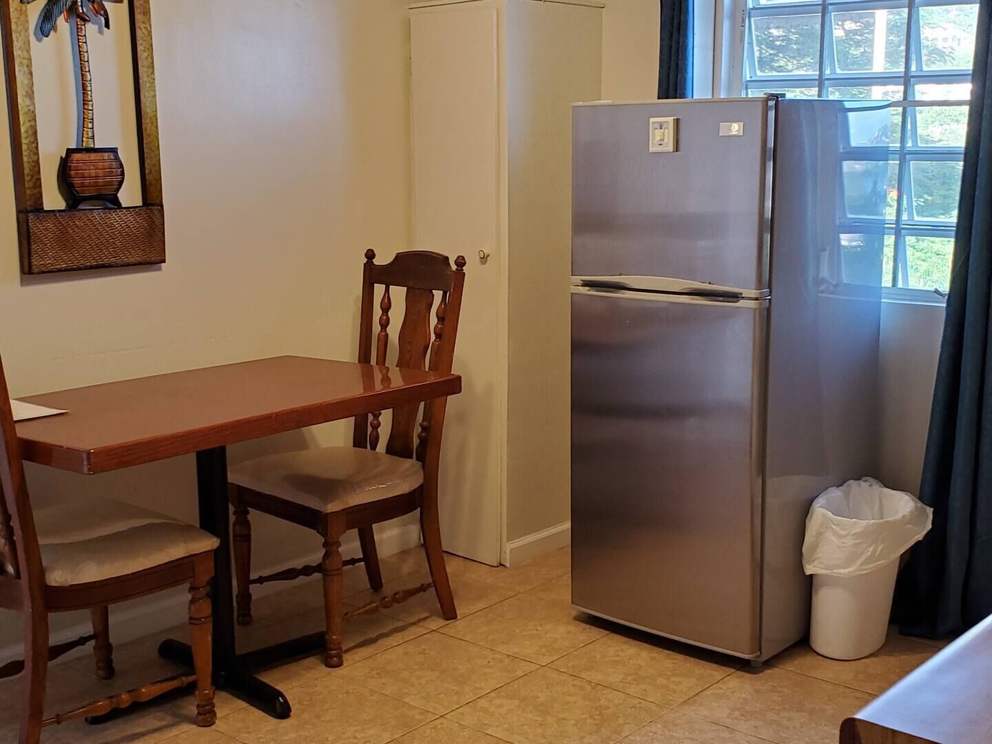 A kitchen with a wooden table and chairs, a stainless steel refrigerator, a trash bin, and a window with curtains.