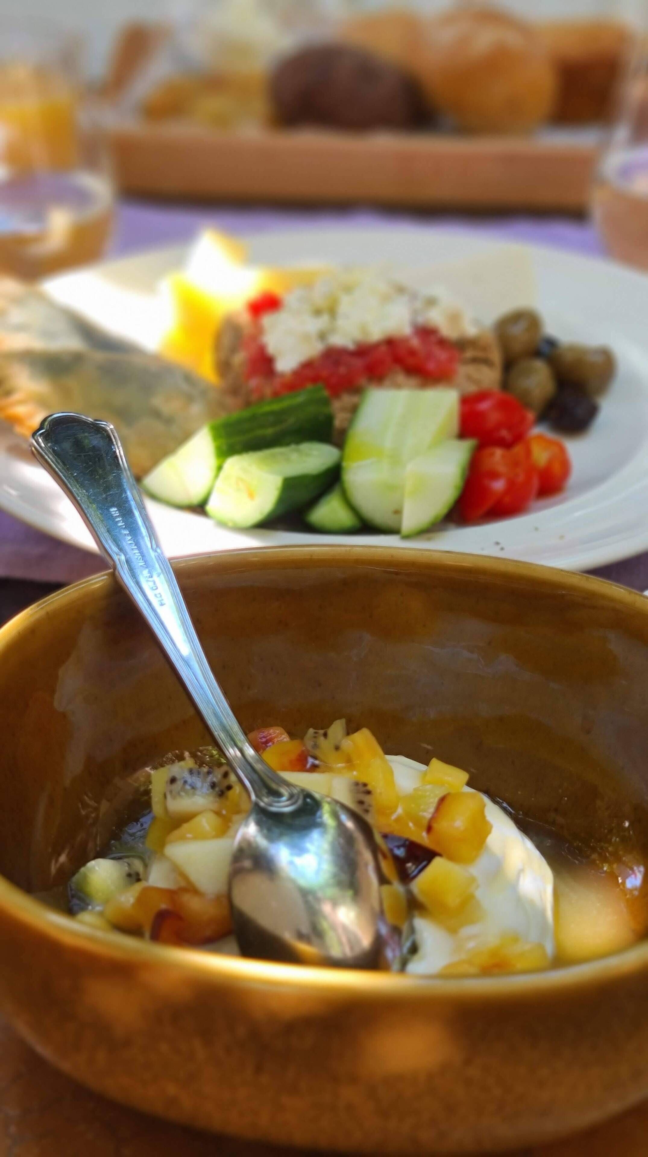 Un saladier rempli de salade de fruits avec une cuillère, une assiette avec des légumes et du fromage, et un panier de pain en arrière-plan.