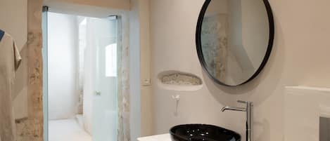 A modern bathroom with a glass shower door, a black basin, and a round mirror on a white countertop.