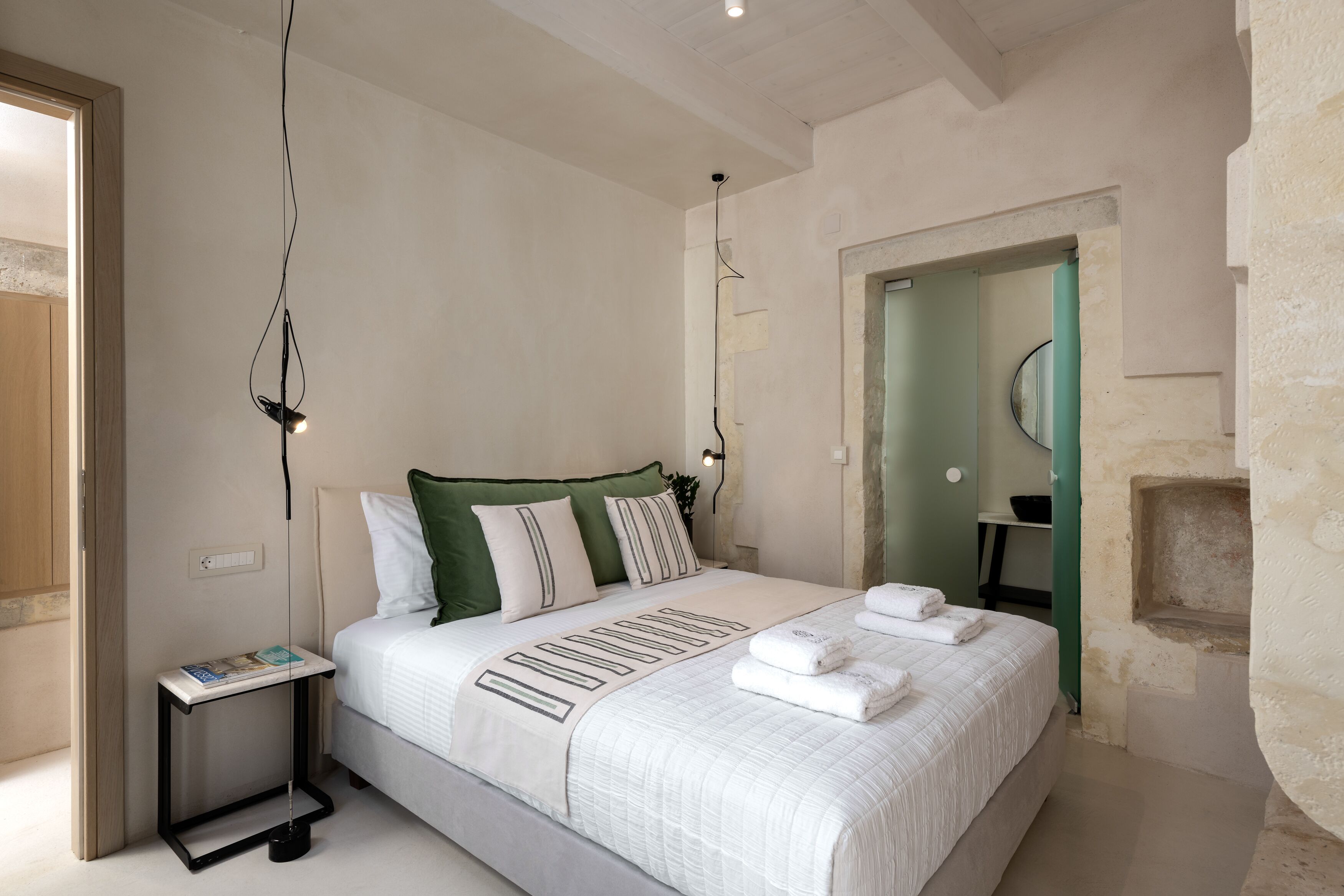 A neatly made bed with a white comforter and green pillows, a bedside table with a magazine, and a wall-mounted light fixture.