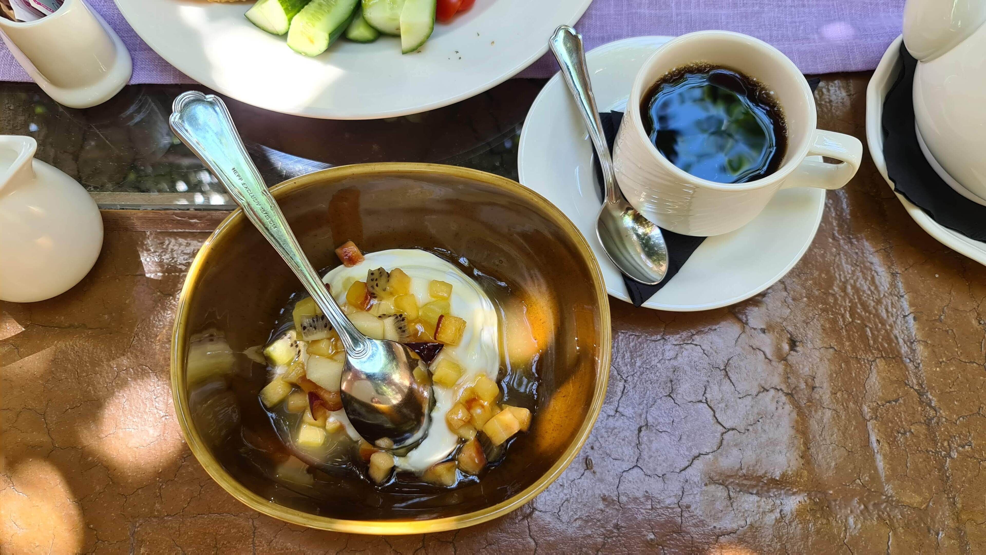 Una mesa con un tazón de ensalada de frutas, una taza de café y un plato de verduras.