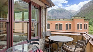 A balcony with a glass table and chairs, overlooking a mountainous landscape and a brick building.