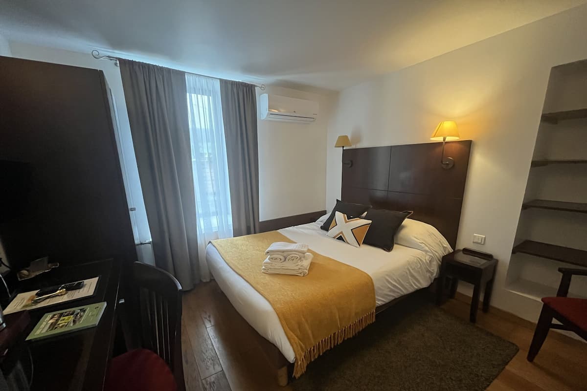 Ein Hotelzimmer mit Bett, Schreibtisch, Stuhl, Lampe und einem fenster mit Vorhängen.