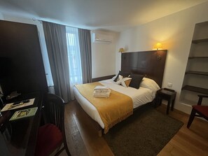 Ein Hotelzimmer mit Bett, Schreibtisch, Stuhl, Lampe und einem fenster mit Vorhängen.
