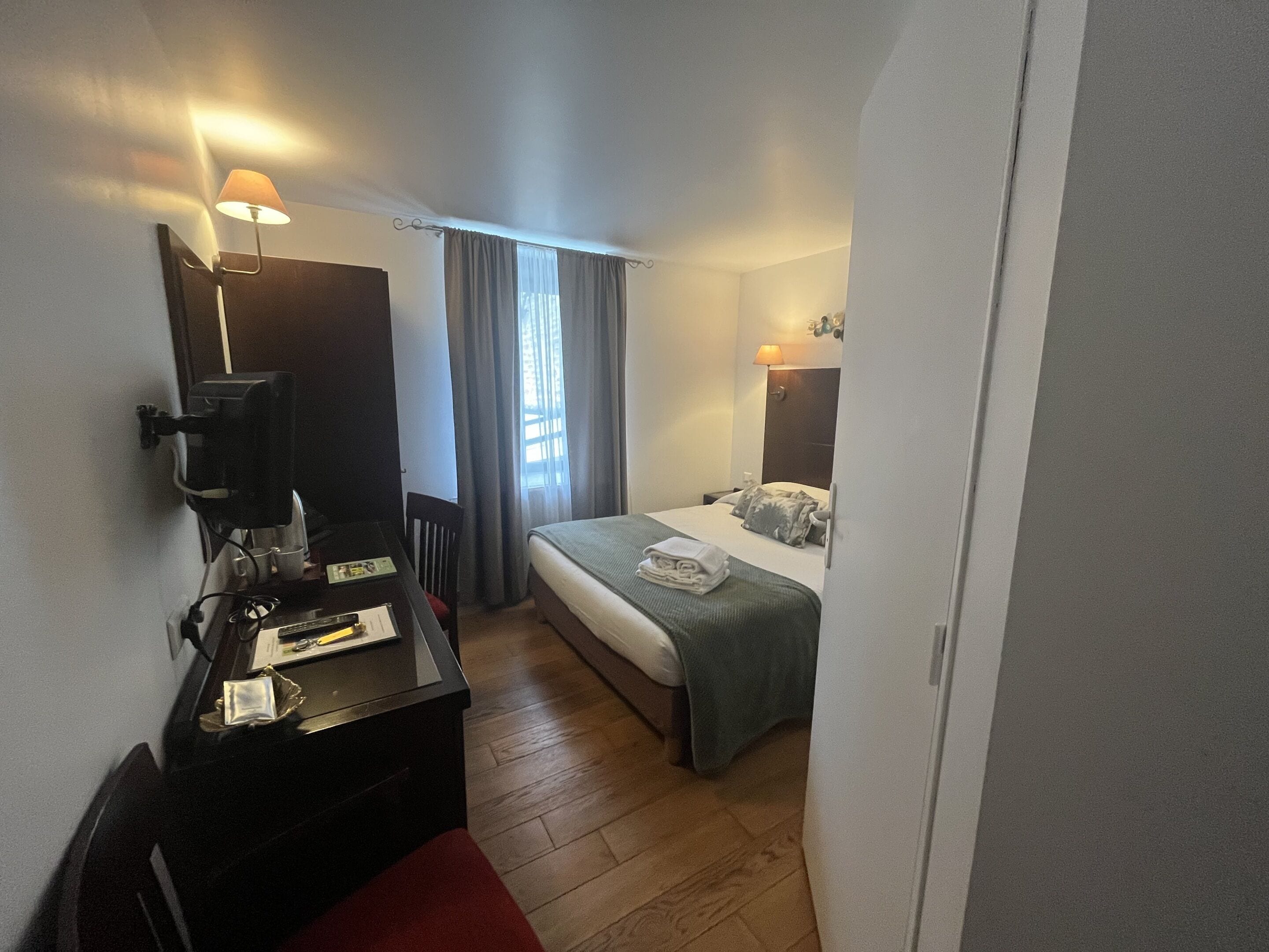 Una habitación de hotel con cama, escritorio con computadora, silla y ventana con cortinas.