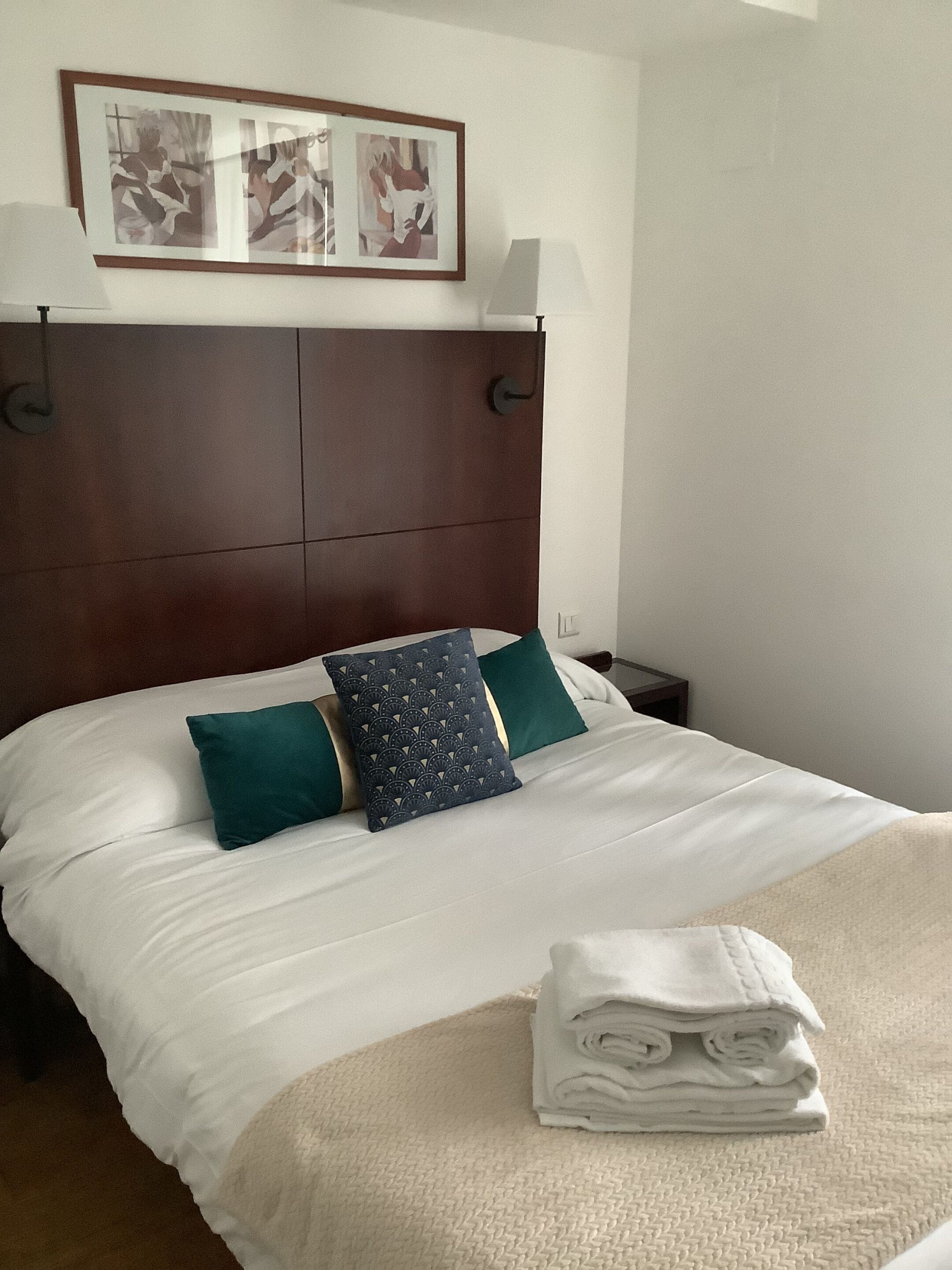 Ein ordentlich gemachtes Bett mit weißen Leinentüchern und türkisfarbenen Kissen, darüber ein gerahmtes Kunstwerk und ein gefaltetes Handtuch auf dem Bett.
