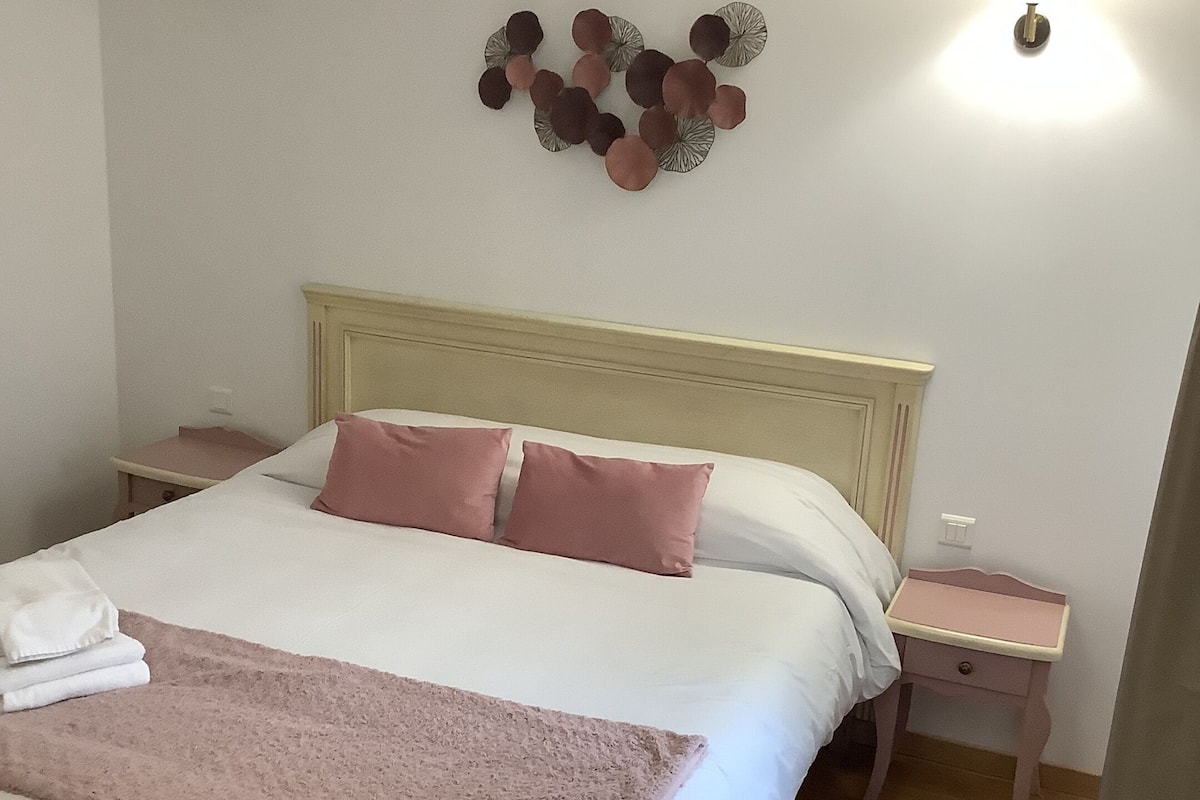 Ein ordentlich bezogenes Bett mit rosa und weißer Bettwäsche, ein hölzerner Kopfteil und ein Nachttisch mit Lampe.