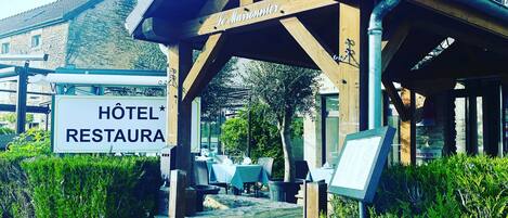 A hotel entrance with a covered patio area, a sign reading "Hôtel Restaura", and a stone wall leading to the entrance.