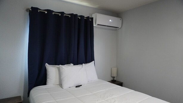 Une chambre d’hôtel avec un lit, du linge de lit blanc, un rideau bleu foncé, une table de chevet avec une lampe et un appareil de climatisation fixé au mur.