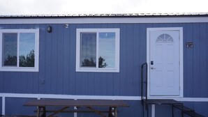 Un bâtiment bleu avec une porte et des fenêtres blanches, une table de pique-nique et une petite table noire placée à côté de la porte.