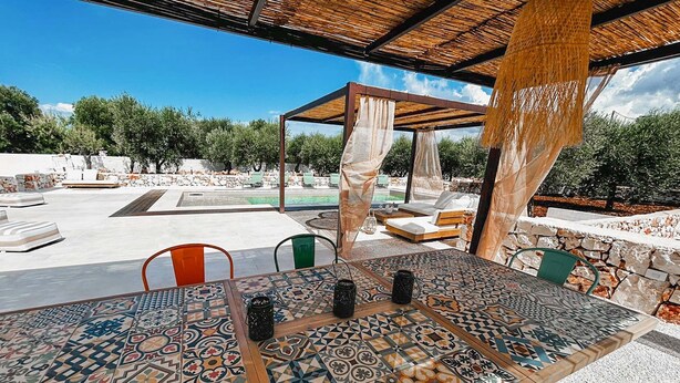 Ein Essbereich im Freien mit einem Tisch und Stühlen mit Muster, eine überdachte Sitzgelegenheit und Blick auf Pool und Grünflächen.