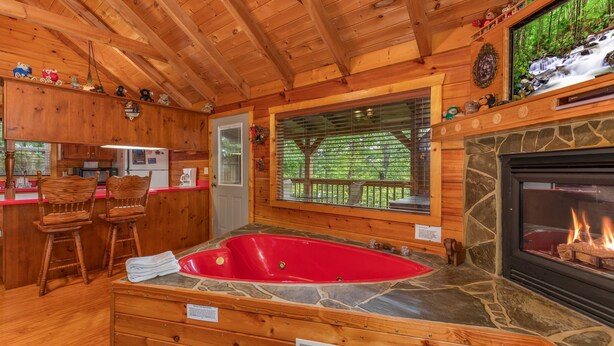 A cozy cabin interior with a stone fireplace, a red hot tub, wooden furniture, and a kitchen area.