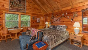 A cozy log cabin bedroom with a bed, a chair, a small table, a lamp, and a window.