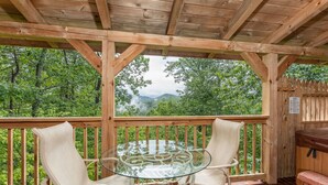 A wooden deck with a glass table and two chairs, overlooking a forested area with mountains in the distance.