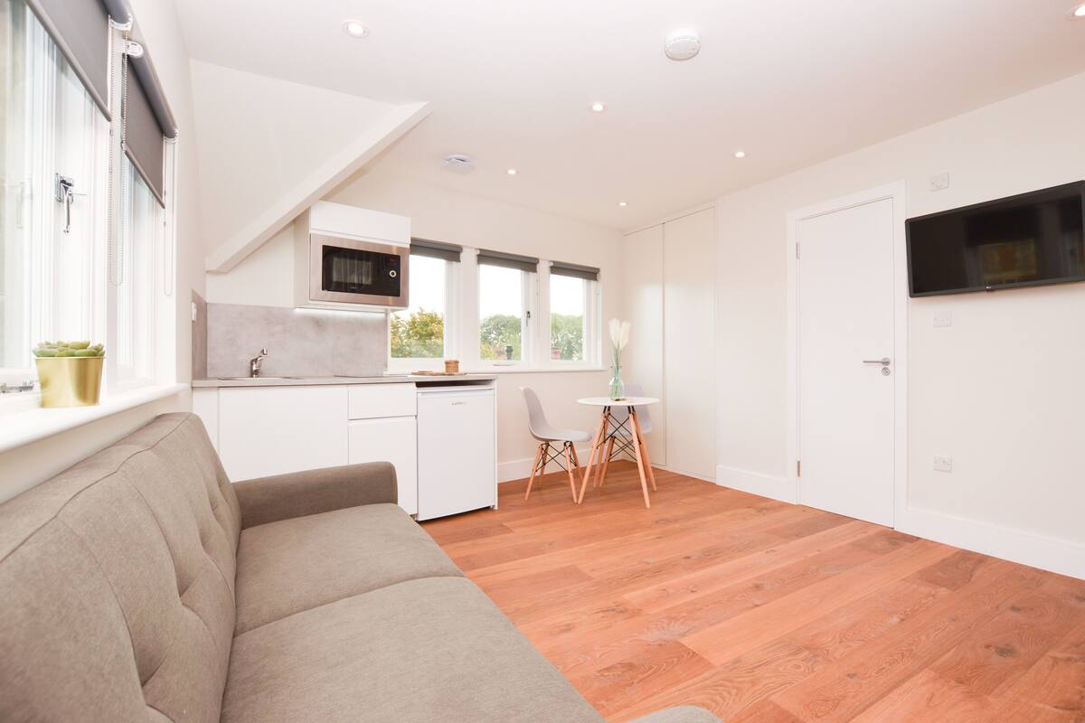 A modern kitchen with white cabinets, a microwave, and a window with blinds. There is a dining area with a table and chairs, and a sofa in the living area.
