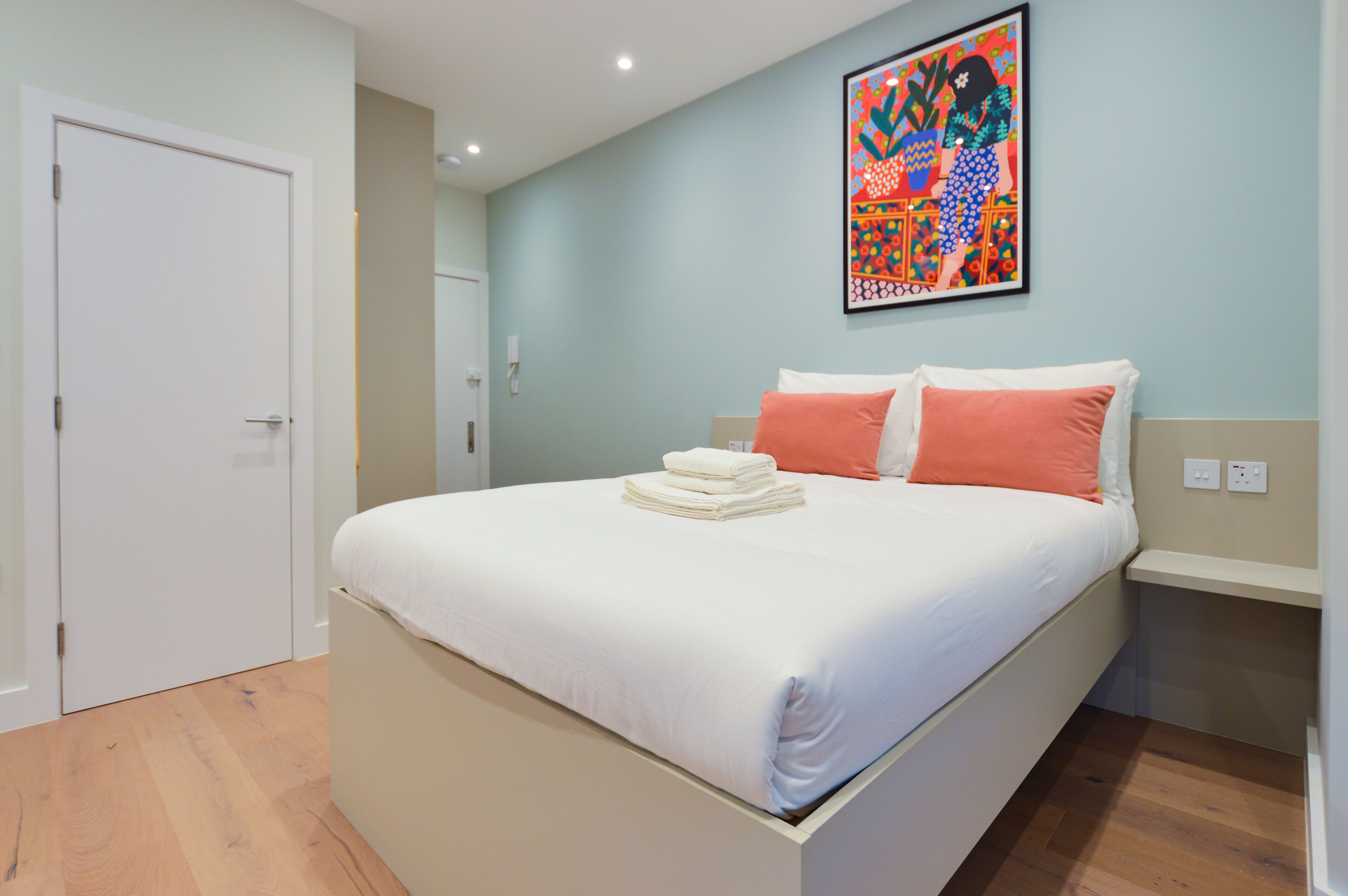 A neatly made bed with white and orange pillows, a colorful abstract painting on the wall, and a white door.