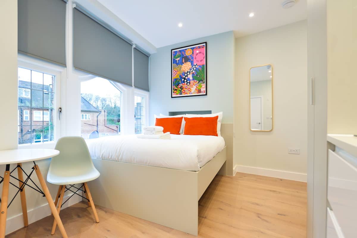A modern bedroom with a bed, a chair, a table, a large window, and a colorful painting on the wall.