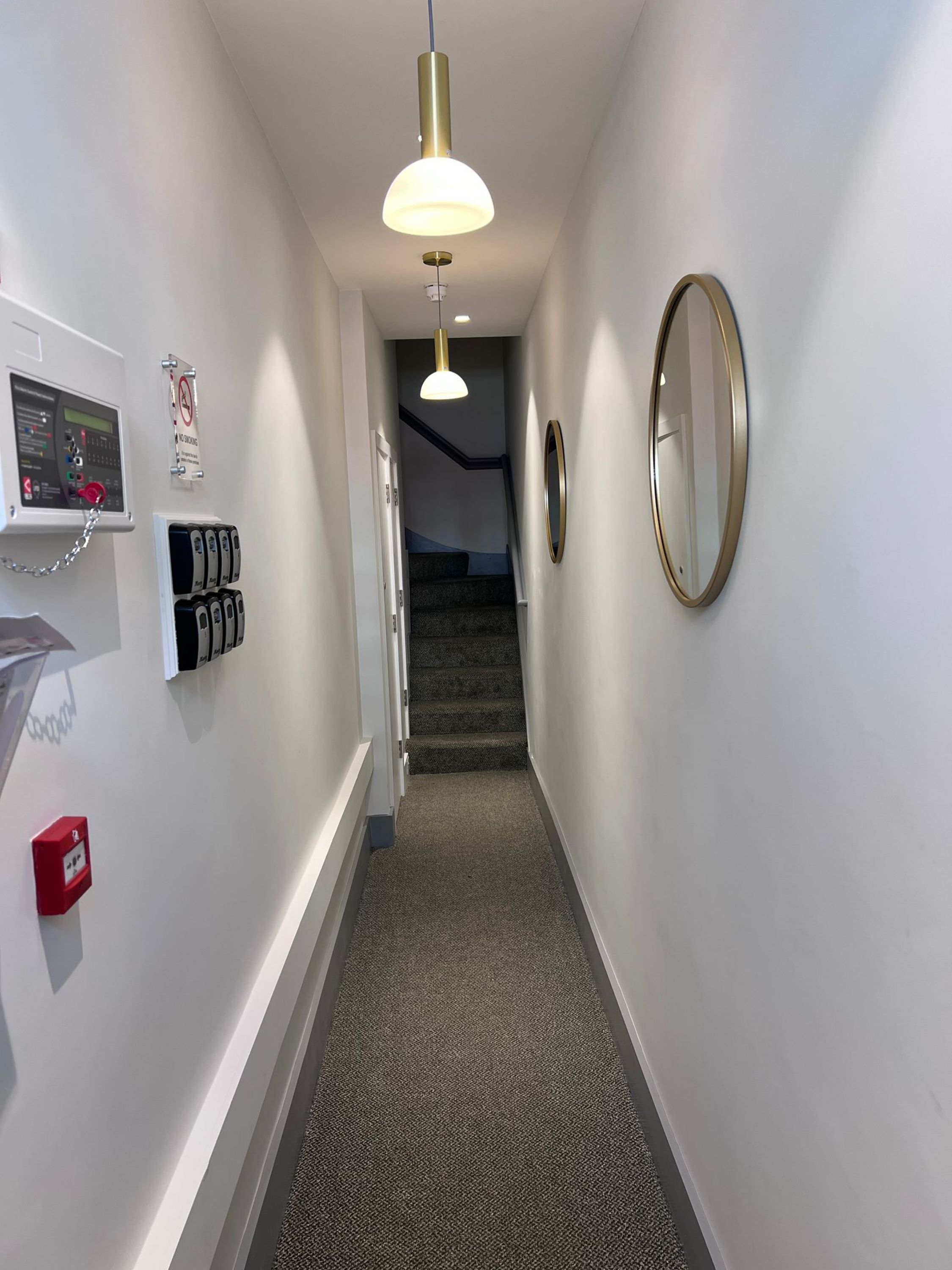 A hallway with a carpeted floor, a staircase, a wall-mounted fire alarm, a smoke detector, a no smoking sign, and a round mirror.