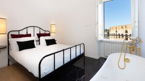 Une chambre à coucher avec un lit, une fenêtre donnant sur des immeubles et une baignoire équipée d’une douchette.