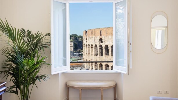 Une pièce avec une fenêtre donnant sur le Colisée, un banc, une plante en pot et un miroir au mur.