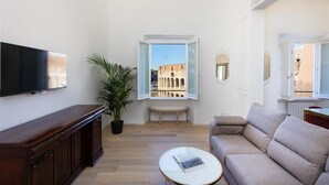 Un salon équipé d’un téléviseur à écran plat, d’un canapé, d’une table basse, d’une plante en pot et d’une fenêtre donnant sur un bâtiment historique.