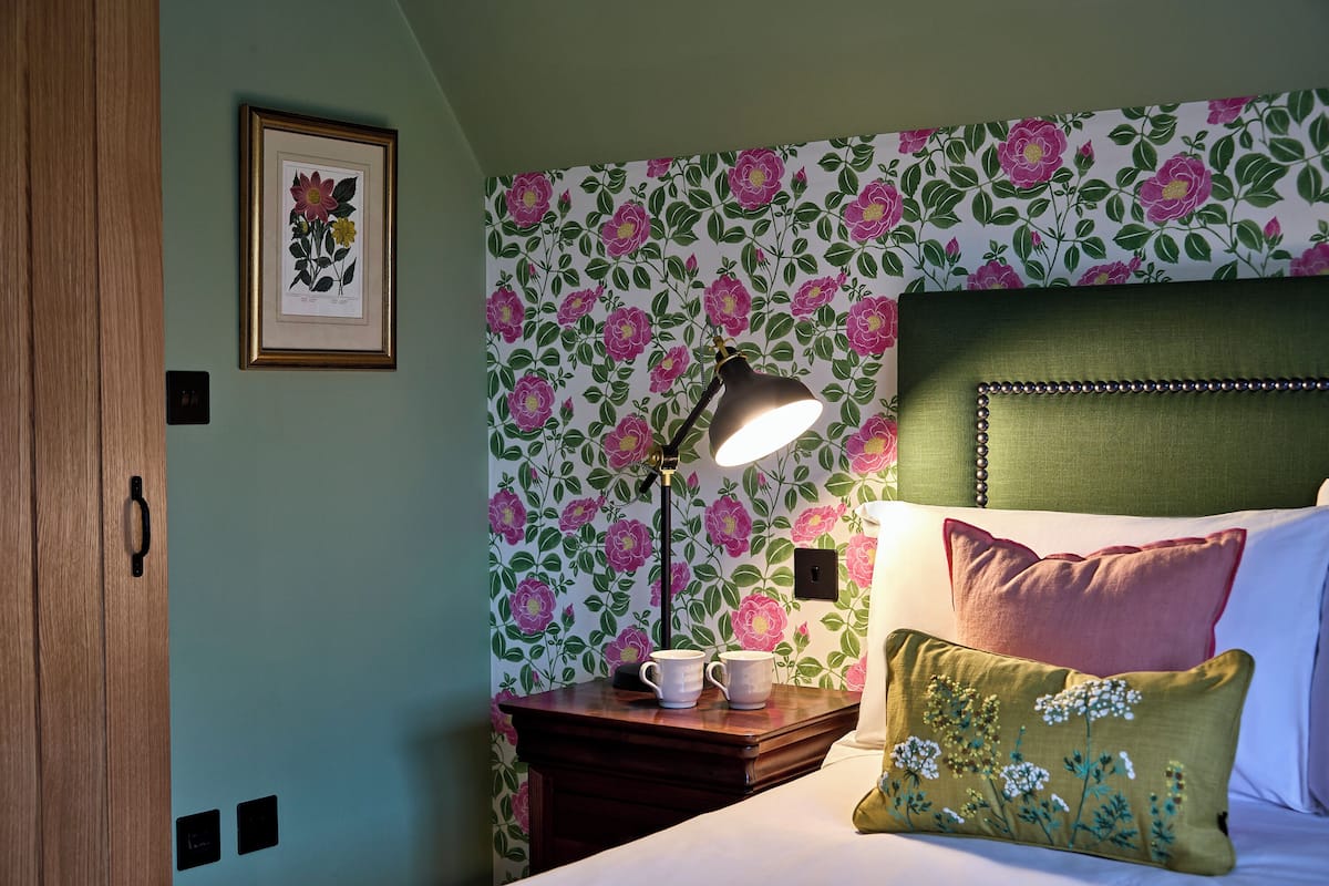 A bedroom with floral wallpaper, a bedside lamp, a framed botanical print, and a bed with pillows.